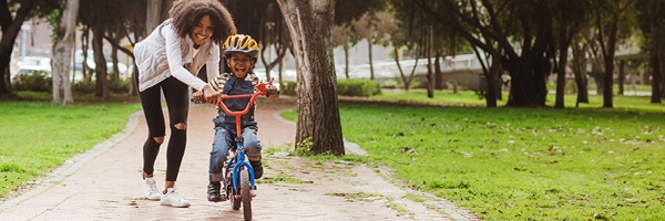 Mother guides her young son who is riding a red bicycle on a brick sidewalk at the park.